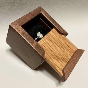 May include: A small wooden ring box featuring a dark brown walnut frame and a sliding light oak lid. The interior is lined with black foam, holding a silver-toned ring with a prominent oval-cut center stone and two smaller marquise-cut side stones.