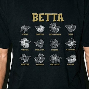 May include: A black short-sleeve t-shirt featuring a gold-colored text graphic that reads BETTA at the top. Below the text are twelve white line-art illustrations of different betta fish tail types, each labeled with its specific name in white text.