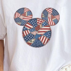 May include: A white short-sleeved t-shirt featuring a central graphic of a mouse head silhouette composed of various patchwork patterns in red, white, and blue, including stars and stripes designs, with a crew neckline and a relaxed fit.