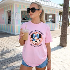 Pink tee with Minnie Mouse in patriotic sunglasses and bow, worn by a woman holding a drink on a boardwalk.