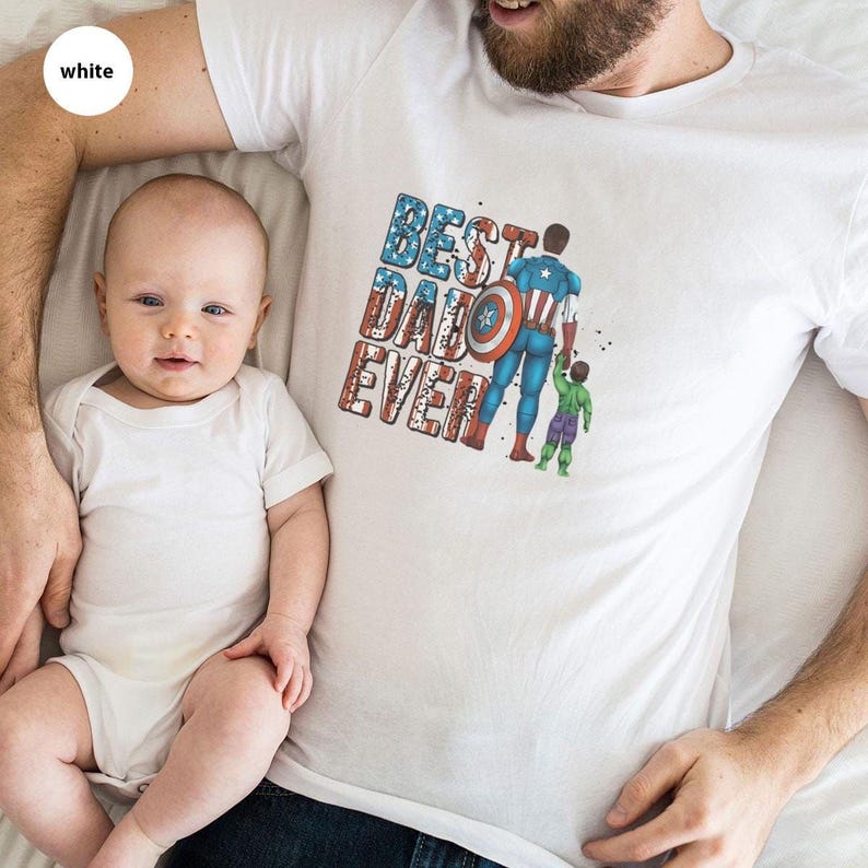 A man and a baby are lying down together, with a t-shirt featuring a graphic design of a superhero character and the text "BEST DAD EVER" printed on it.