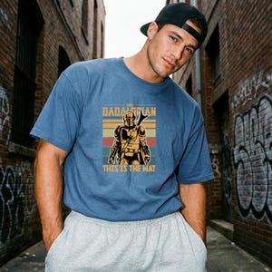 A man wearing a blue t-shirt with a graphic of a character from the Star Wars franchise, specifically The Mandalorian, standing in a graffiti-covered alleyway.