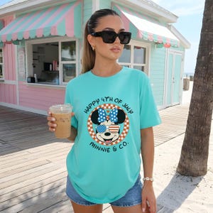 Aqua t-shirt with "Happy 4th of July Minnie & Co." design, worn by a woman holding a drink outdoors.