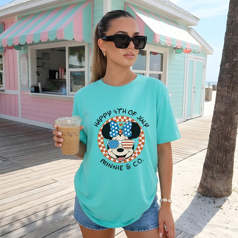 Aqua t-shirt with "Happy 4th of July Minnie & Co." design, worn by a woman holding a drink outdoors.