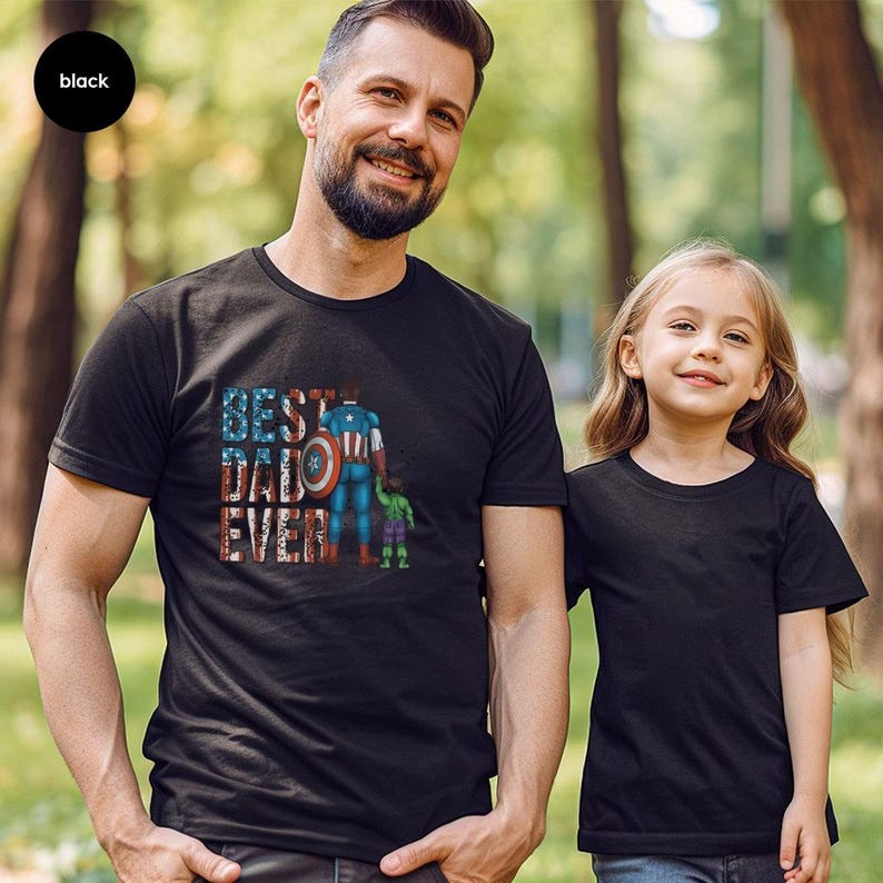 A man and a young girl are standing together in a park, both wearing black t-shirts with a graphic design.