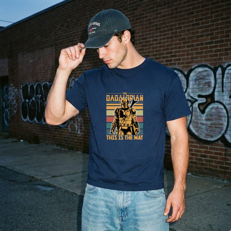 A man wearing a blue t-shirt with a graphic design and a baseball cap stands in front of a brick wall with graffiti.