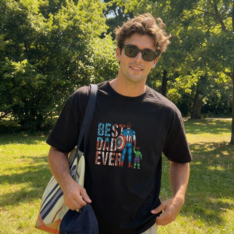 A man wearing a black t-shirt with a graphic design of a superhero character and the text "Best Dad Ever" is standing in a grassy area surrounded by trees.