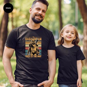 A man and a young girl are standing together in a park, both wearing black t-shirts with a graphic design featuring a character from the Star Wars franchise.