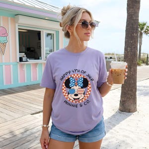 Blonde woman in sunglasses wearing a lavender Minnie Mouse "Happy 4th of July" tee, holding an iced coffee.