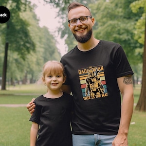 A man and a young girl wearing matching black t-shirts with a graphic design, standing together in a park.