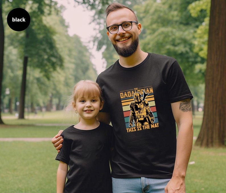 A man and a young girl wearing matching black t-shirts with a graphic design, standing together in a park.