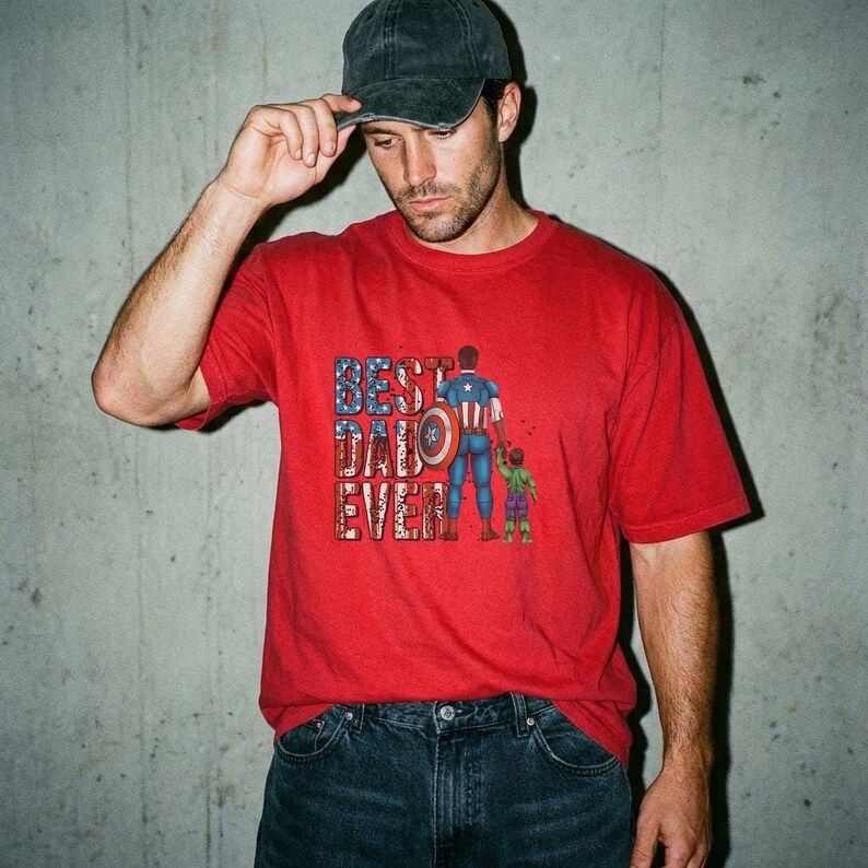 A man wearing a red t-shirt with a graphic design of a superhero and the text "Best Dad Ever" is standing in front of a concrete wall.