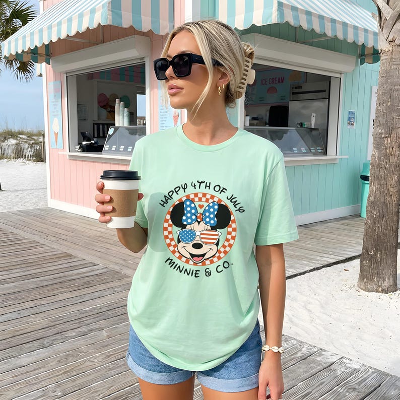 Light green tee with Minnie Mouse in patriotic sunglasses, worn by a woman on a boardwalk.