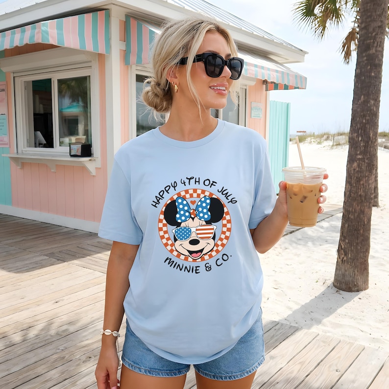 Light blue Minnie Mouse 4th of July tee worn by smiling girl at beachside stand.