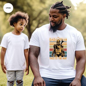 A young child and an adult man wearing a white t-shirt with a graphic design, standing together in a grassy outdoor setting.