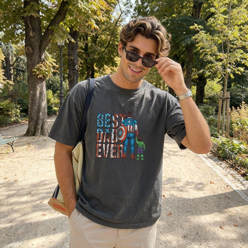 A man wearing a gray t-shirt with a graphic design and sunglasses is standing in a park surrounded by trees.