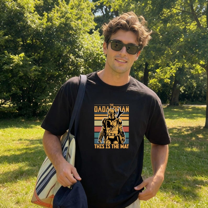 A man wearing a black t-shirt with a graphic design and sunglasses stands in a grassy area with trees in the background.