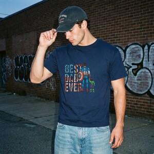 A man wearing a blue t-shirt with a graphic design and a baseball cap stands in front of a brick wall with graffiti.
