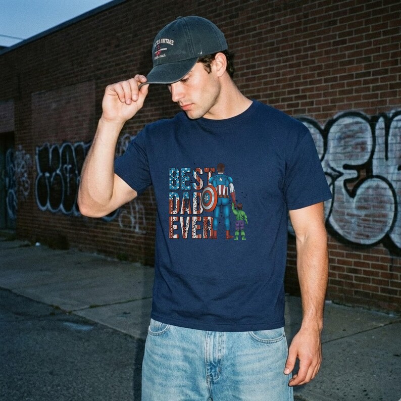 A man wearing a blue t-shirt with a graphic design and a baseball cap stands in front of a brick wall with graffiti.
