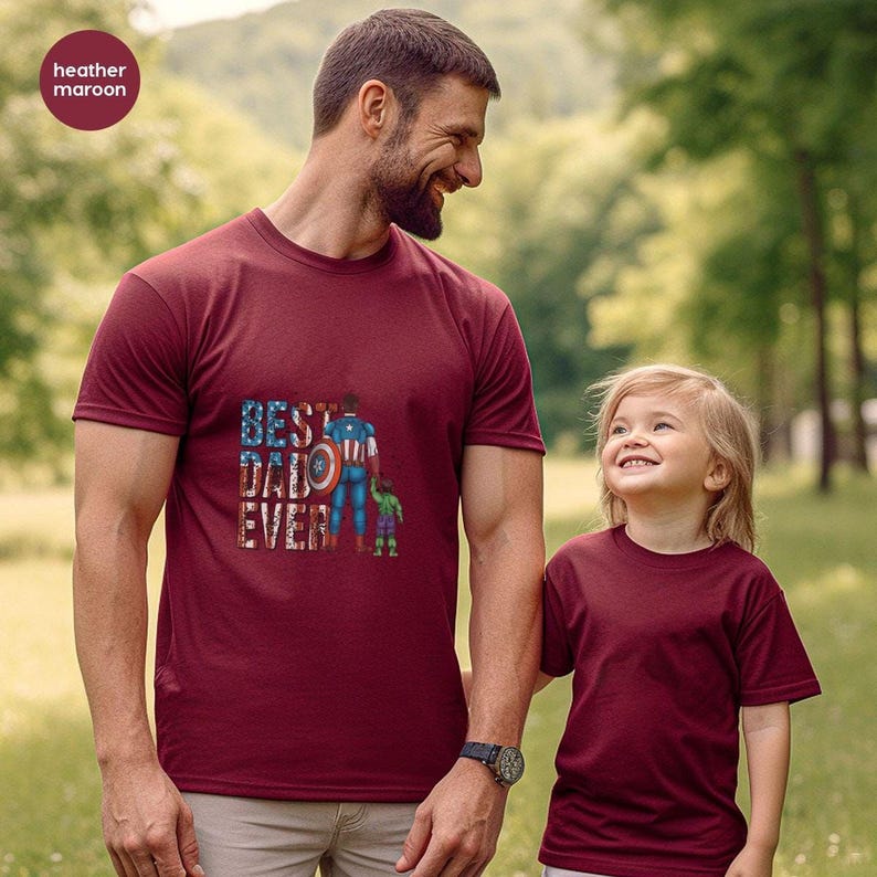 A man and a young girl wearing matching maroon t-shirts with a graphic design, standing together in a park.