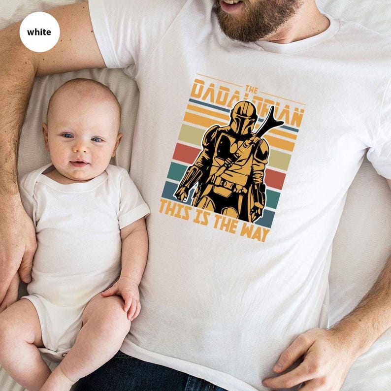 A man and a baby are lying down together, with the man wearing a white t-shirt featuring a graphic of a character from the Star Wars franchise.
