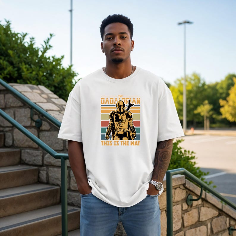 A man wearing a white t-shirt with a graphic design of a character from the Star Wars franchise, specifically The Mandalorian, standing on a set of stairs.