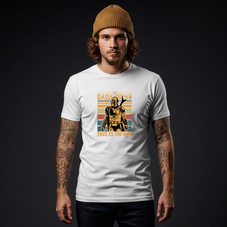 A man wearing a white t-shirt with a graphic design of a character from the Star Wars franchise, specifically the Mandalorian, standing against a dark background.