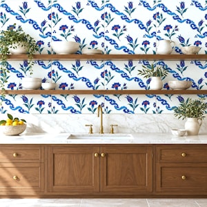 May include: A kitchen featuring wooden cabinets, a white marble countertop, and a sink with a brass faucet. The wall is covered in white tiles with a repeating blue and green floral pattern. Two wooden shelves hold various ceramic bowls and plants.