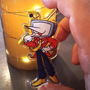May include: A clear acrylic keychain featuring a cartoon character with a television set for a head, a long pointed nose, a red blazer, a yellow tie, dark blue trousers, and yellow shoes, with a small loop at the top for attachment.