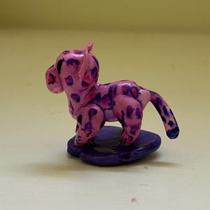 May include: A handmade pink and purple leopard figurine. The small sculpture features a spotted pattern in darker purple and is posed on a purple base. The figurine has a playful, cartoonish style.