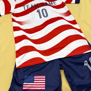 May include: A short-sleeve athletic jersey and shorts set featuring a red and white wavy stripe pattern. The jersey displays the text TEAM NAME, the number 10, and a small American flag. The navy blue shorts feature a large American flag graphic.