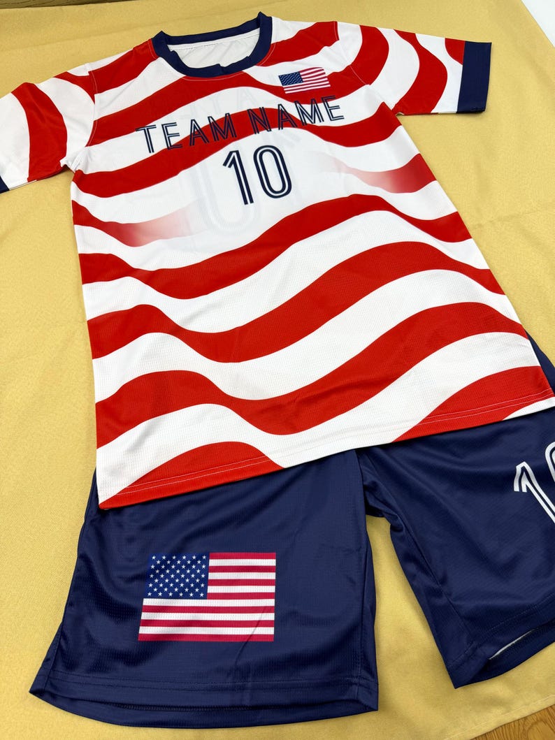 May include: A short-sleeve athletic jersey and shorts set featuring a red and white wavy stripe pattern. The jersey displays the text TEAM NAME, the number 10, and a small American flag. The navy blue shorts feature a large American flag graphic.