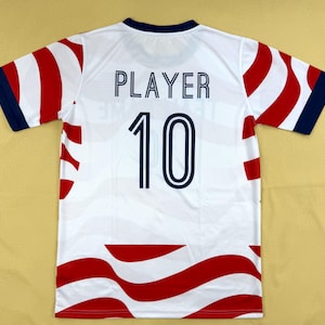 May include: A white short-sleeved sports jersey featuring a wavy red pattern across the body and sleeves. The back displays the word PLAYER in dark blue capital letters above the number 10 in a matching dark blue font with a white outline.