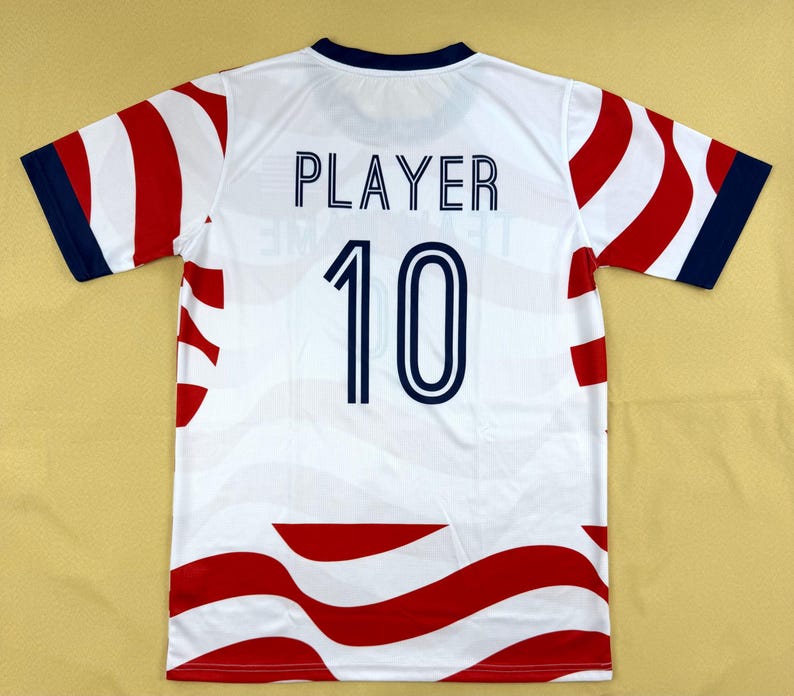 May include: A white short-sleeved sports jersey featuring a wavy red pattern across the body and sleeves. The back displays the word PLAYER in dark blue capital letters above the number 10 in a matching dark blue font with a white outline.