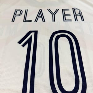 May include: A white sports jersey featuring a dark navy blue collar and the word PLAYER printed in dark navy blue block letters above the large dark navy blue number 10. The fabric has a subtle textured mesh pattern throughout the garment.