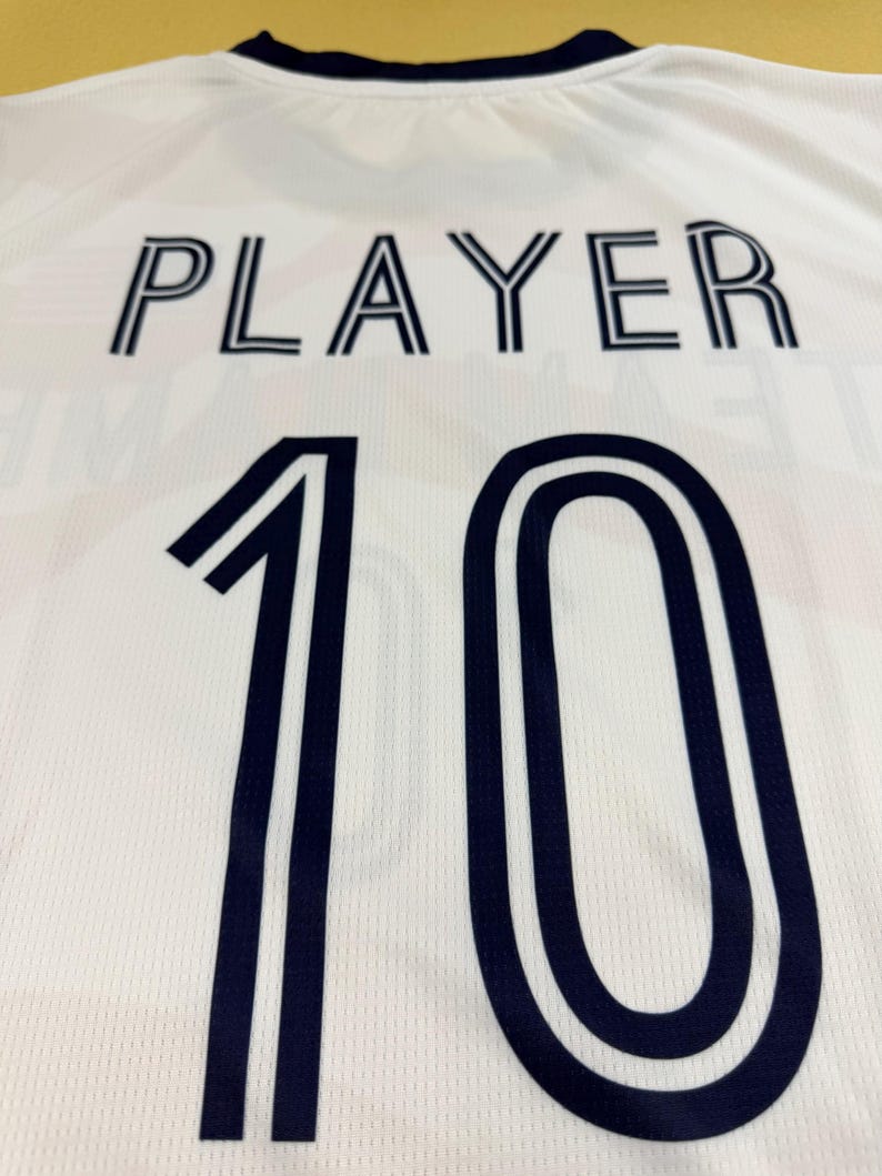 May include: A white sports jersey featuring a dark navy blue collar and the word PLAYER printed in dark navy blue block letters above the large dark navy blue number 10. The fabric has a subtle textured mesh pattern throughout the garment.