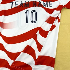 May include: A short-sleeved jersey featuring a wavy red and white striped pattern. The shirt includes a dark blue crew neckline, a small American flag graphic, the text "TEAM NAME" in dark blue, and the number "10" printed in dark blue.