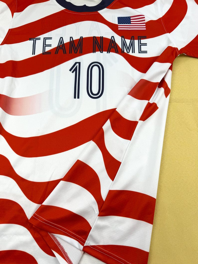 May include: A short-sleeved jersey featuring a wavy red and white striped pattern. The shirt includes a dark blue crew neckline, a small American flag graphic, the text "TEAM NAME" in dark blue, and the number "10" printed in dark blue.