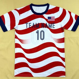 May include: A short-sleeve t-shirt featuring a wavy red and white striped pattern. The shirt has a dark blue crew neckline and sleeve cuffs. The text "TEAM NAME" is printed in dark blue, with the number "10" below it and a small US flag icon.