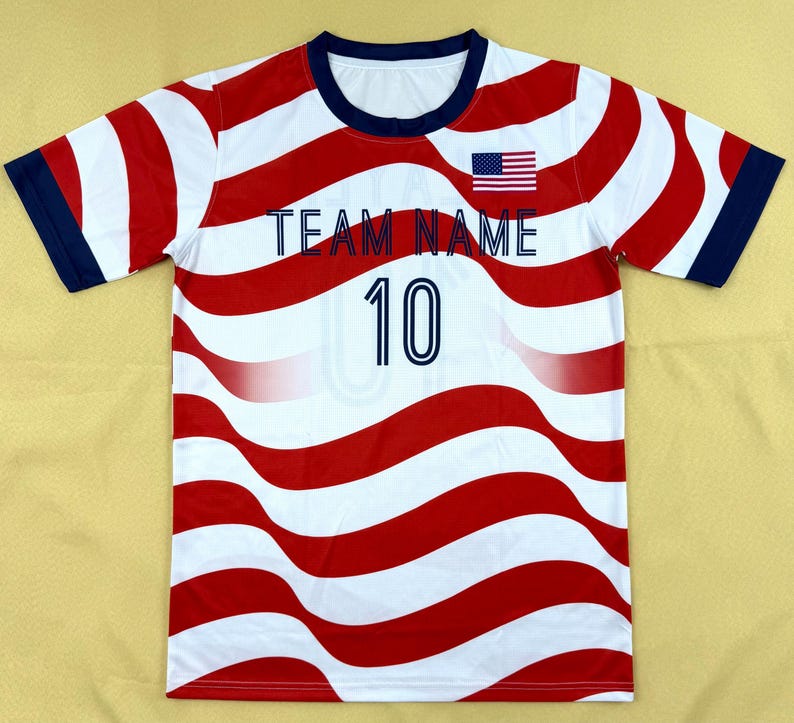 May include: A short-sleeve t-shirt featuring a wavy red and white striped pattern. The shirt has a dark blue crew neckline and sleeve cuffs. The text "TEAM NAME" is printed in dark blue, with the number "10" below it and a small US flag icon.