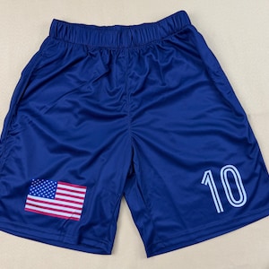 May include: Dark blue athletic shorts featuring an elastic waistband, a printed United States flag on the lower left leg, and the number 10 in white outlined font on the lower right leg. The shorts are made of a smooth, lightweight fabric.