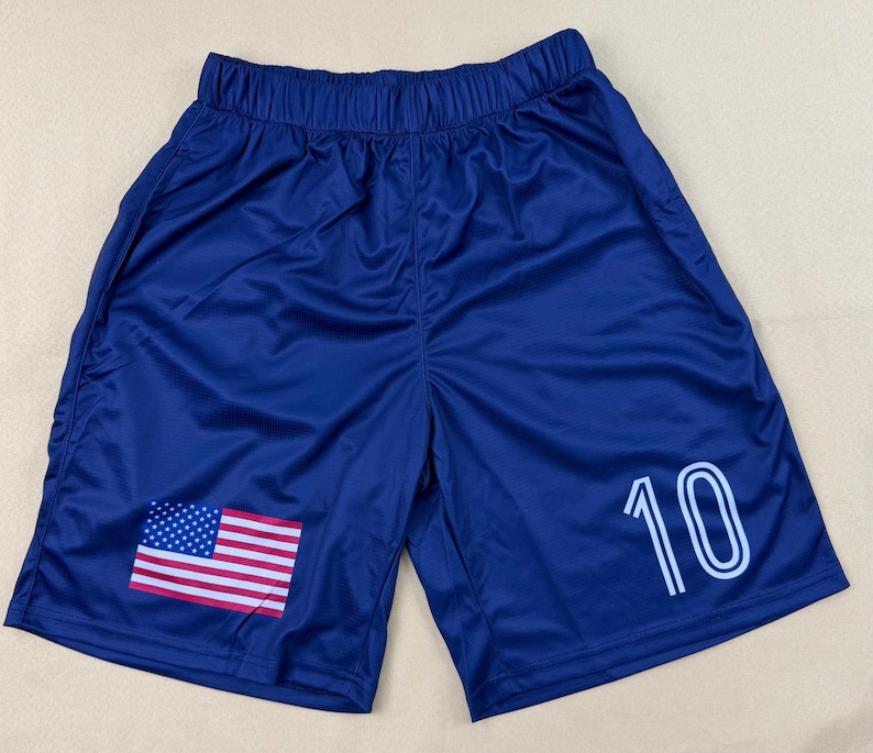 May include: Dark blue athletic shorts featuring an elastic waistband, a printed United States flag on the lower left leg, and the number 10 in white outlined font on the lower right leg. The shorts are made of a smooth, lightweight fabric.