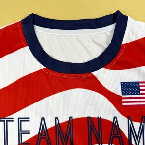 May include: A white athletic jersey featuring a red and white wavy stripe pattern, a dark blue crew neckline, and a small United States flag graphic. The text "TEAM NAME" is printed in dark blue capital letters across the chest area.