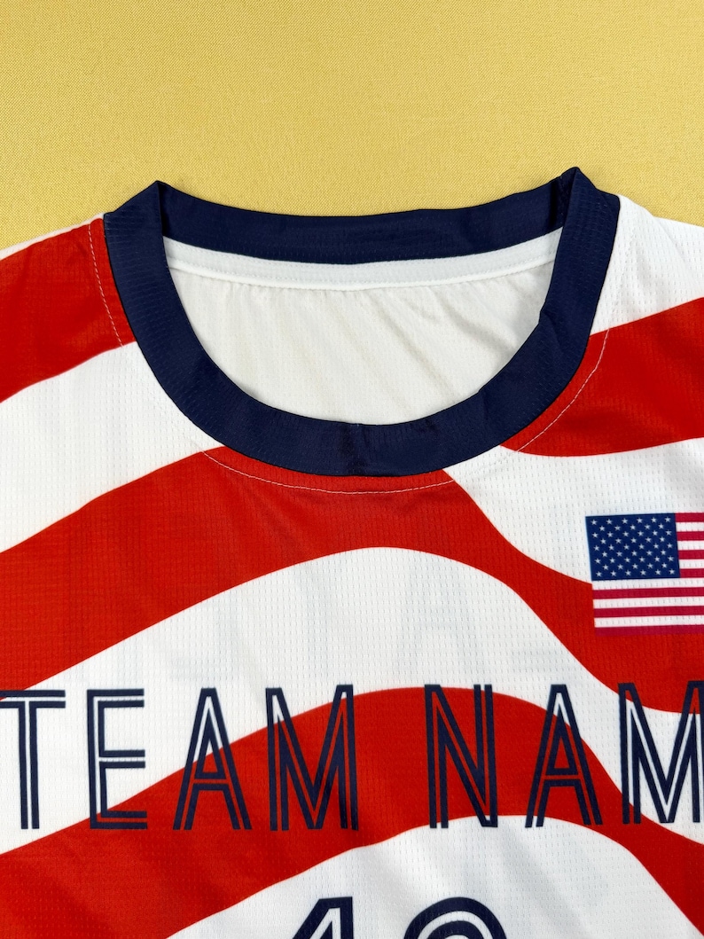 May include: A white athletic jersey featuring a red and white wavy stripe pattern, a dark blue crew neckline, and a small United States flag graphic. The text "TEAM NAME" is printed in dark blue capital letters across the chest area.