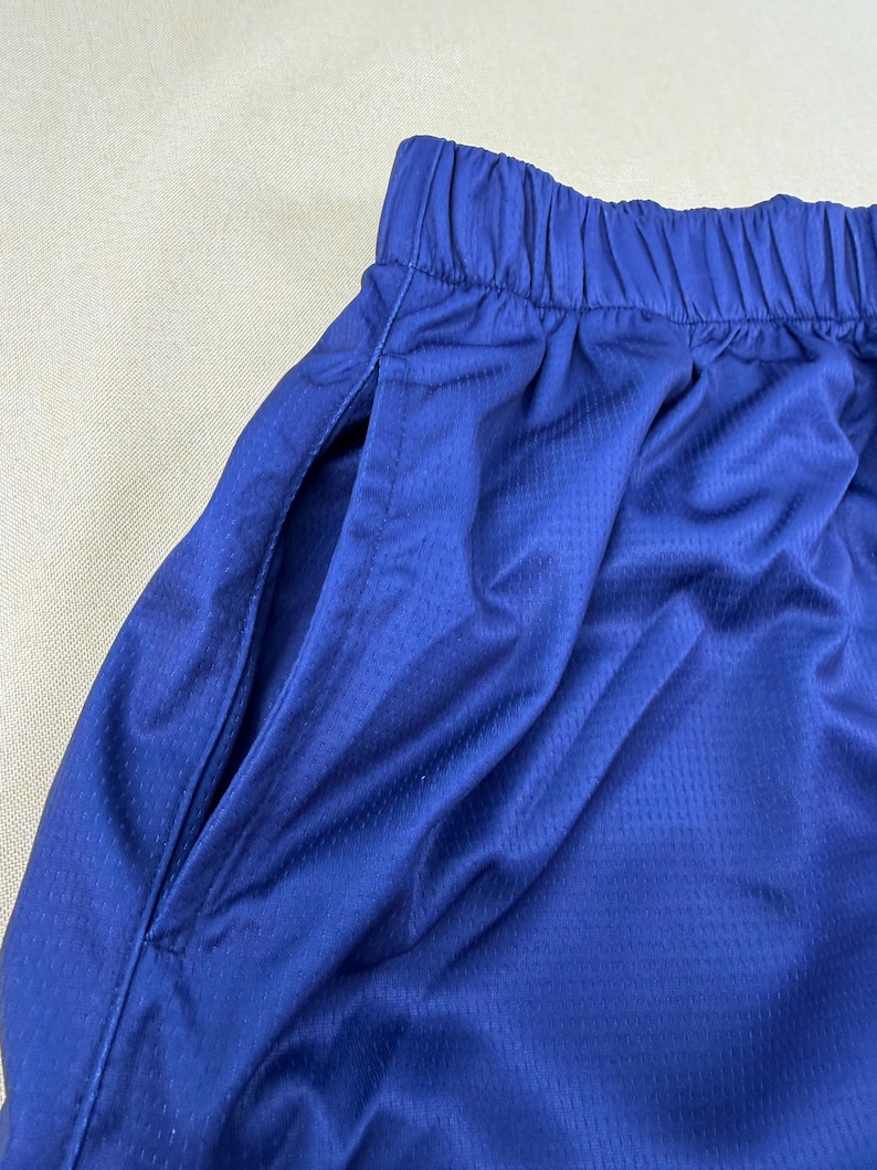 May include: A pair of royal blue athletic shorts featuring a textured, breathable mesh fabric. The shorts have an elasticized waistband with gathered fabric and a deep side pocket opening visible on the left side of the garment.