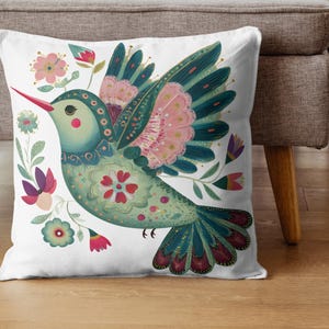 May include: White square throw pillow featuring a colorful illustration of a stylized bird with floral accents. The bird has teal, pink, and red details. The pillow is on a wooden floor next to a gray couch.
