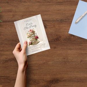 May include: A hand holds a tea party invitation with floral and teacup illustrations. The invitation reads "Eve's Tea Party" with event details. A blue notebook with a pen, eyeglasses, and greenery are also on the wooden surface.