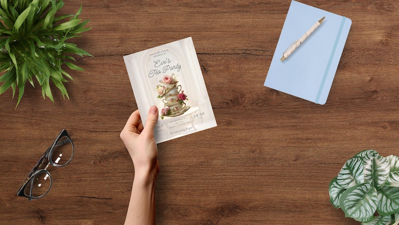 May include: A hand holds a tea party invitation with floral and teacup illustrations. The invitation reads "Eve's Tea Party" with event details. A blue notebook with a pen, eyeglasses, and greenery are also on the wooden surface.