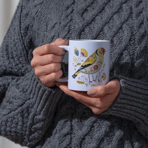 May include: White ceramic mug featuring a colorful illustration of a bird with yellow, red, and black details, surrounded by floral accents. The mug is held by a person wearing a gray cable-knit sweater. The mug is a standard 11 oz size.