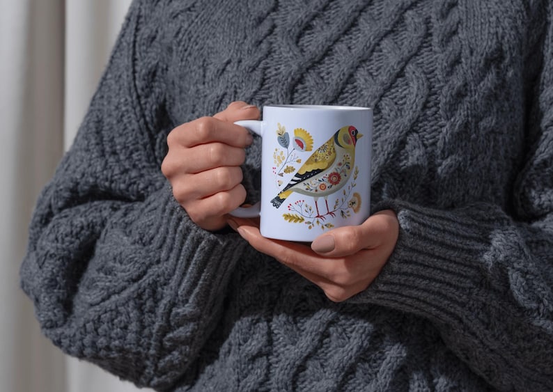 May include: White ceramic mug featuring a colorful illustration of a bird with yellow, red, and black details, surrounded by floral accents. The mug is held by a person wearing a gray cable-knit sweater. The mug is a standard 11 oz size.